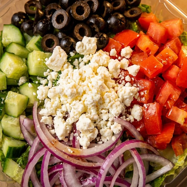 Fresh Greek salad with cucumbers, olives, feta, tomatoes, and red onions in a plastic container.