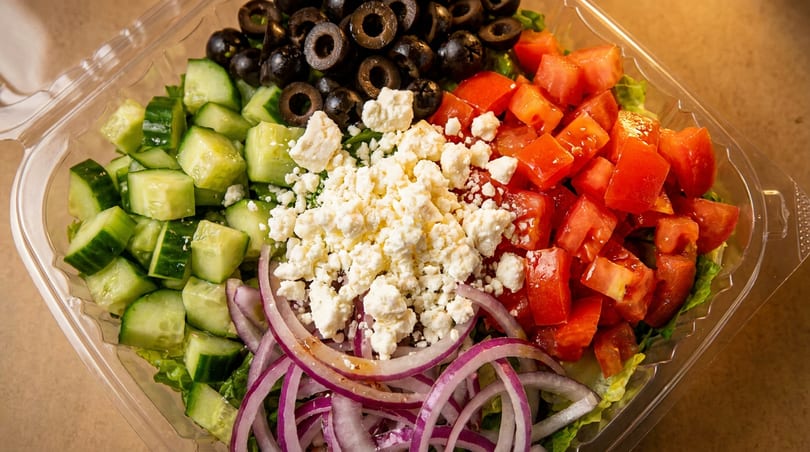 Fresh Greek salad with cucumbers, olives, feta, tomatoes, and red onions in a plastic container.