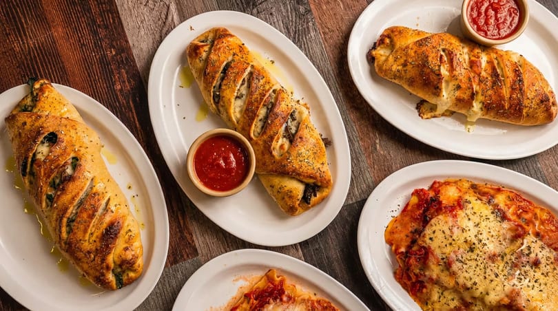 Multiple golden-brown strombolis and cheesy lasagna dishes with marinara sauce on a rustic wooden table.