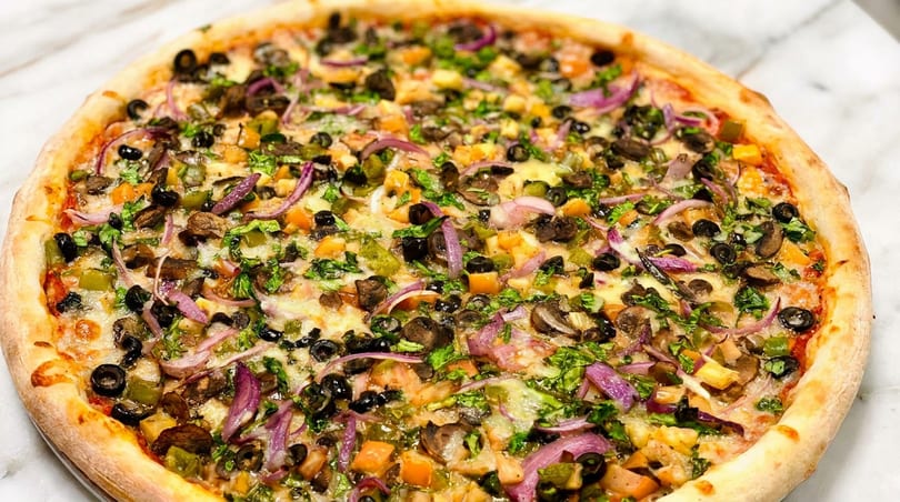 Whole vegetarian pizza topped with olives, red onions, mushrooms, bell peppers, and herbs on a white surface.